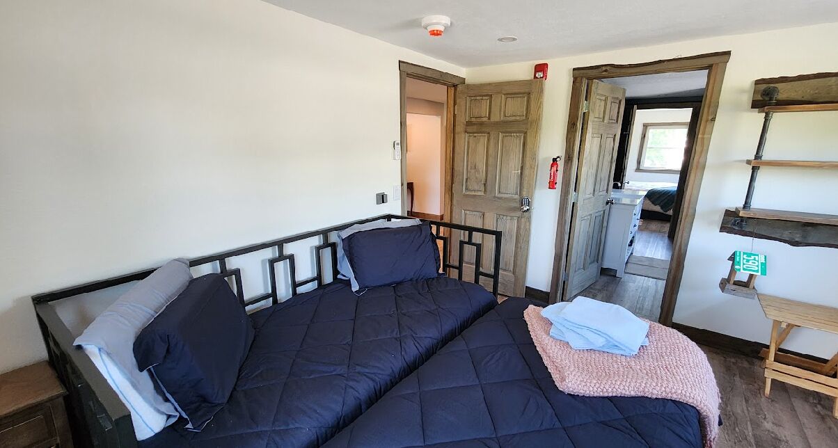 Un lit simple avec un cadre noir et une literie sombre, une porte en bois et une petite table en bois sur laquelle est posée une enseigne verte.
