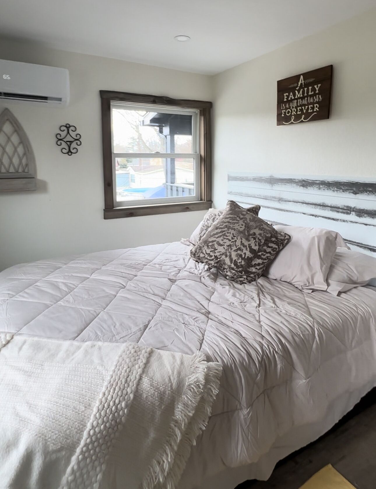 A bedroom with a bed, a window, a wall-mounted air conditioner, and a decorative sign.