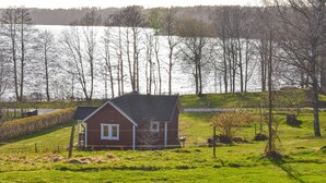 Een klein rood huis met een schuin dak, omgeven door een groen veld en bomen, in de buurt van een waterpartij.