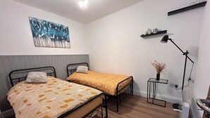 Una habitación pequeña y bien iluminada con dos camas, un escritorio, una silla, una lámpara y un cuadro en la pared.
