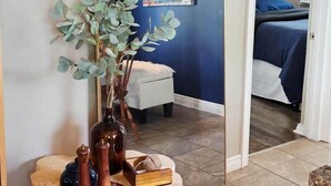 A room with a mirror reflecting a bed, a wall with a colorful poster, and a plant on a wooden stand.