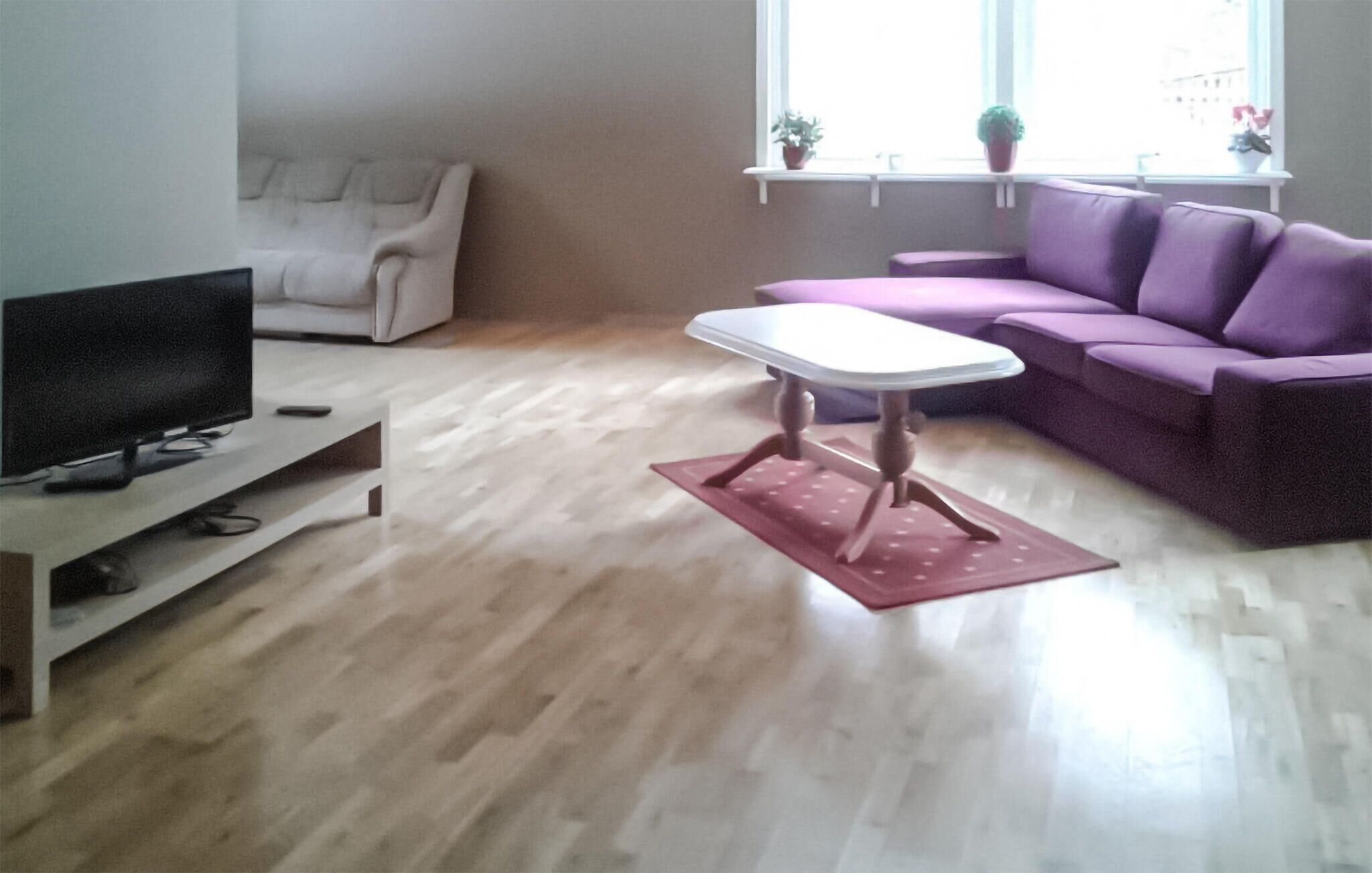 Un salon avec un téléviseur posé sur un meuble, un canapé blanc, un canapé violet et une petite table sur laquelle est posé un tapis rouge.
