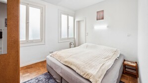 A bedroom with a bed, two windows, a heater, and a small wooden bench.