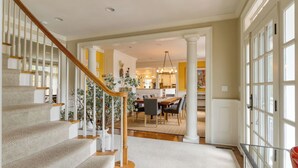 A staircase with wooden railings, a dining area with a table and chairs, and a kitchen visible through an open doorway. - Secluded Estate Optional Guest House, Pond, Playroom, Gym & Theater Sunny Pond by Avantstay (Austerlitz)