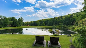 A serene lakeside setting with two lounge chairs facing the water, surrounded by lush greenery and a clear sky.