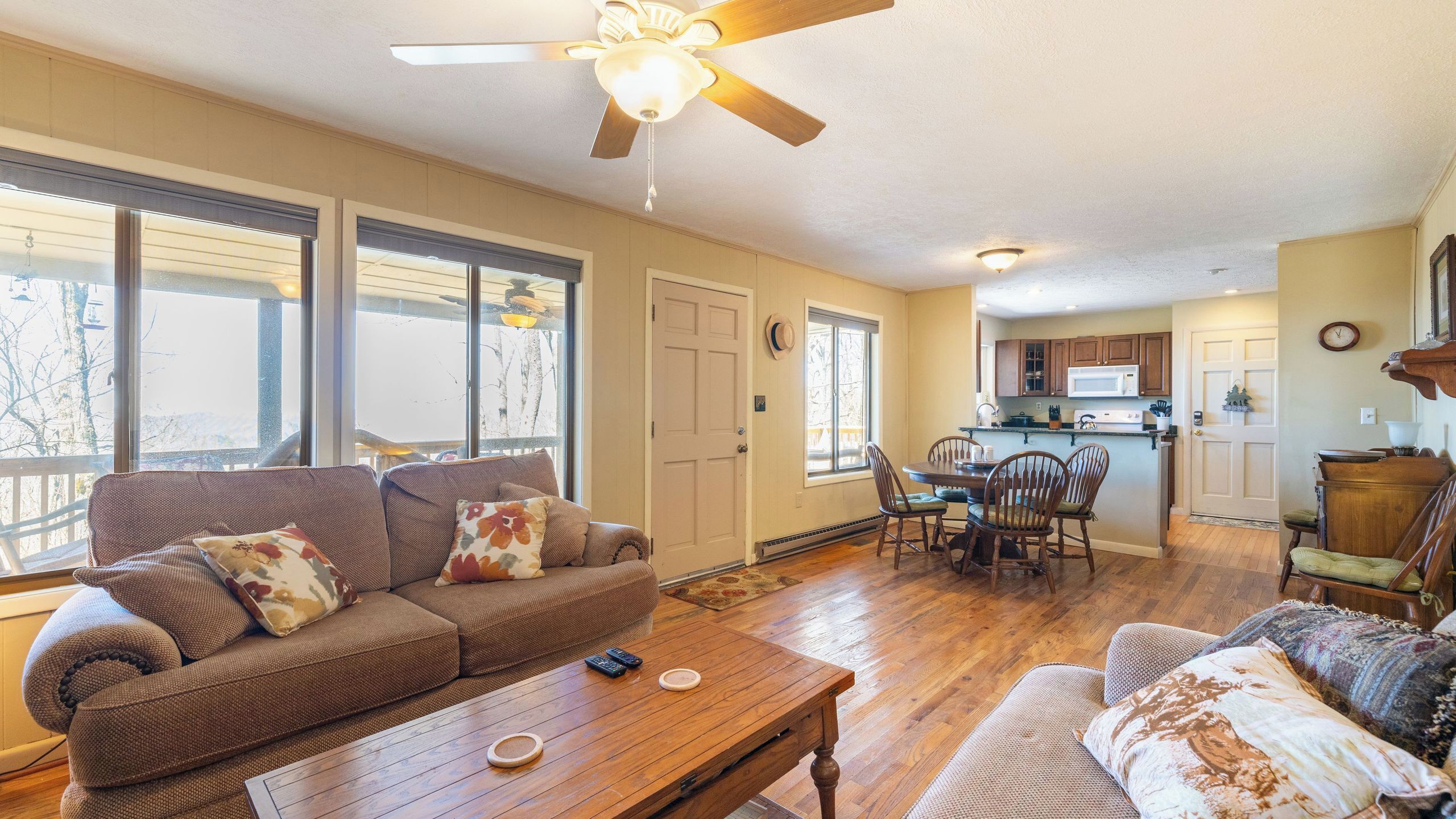 A spacious living room with a sofa, coffee table, and dining area with chairs. A ceiling fan and kitchen are visible in the background.