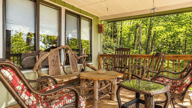 A covered outdoor seating area with wicker furniture and a glass table, overlooking a forested area. - Ash Vale by Avantstay Expansive Deck + Ping Pong + Covered Porch Blue Ridge Mountain Escape (Mars Hill)