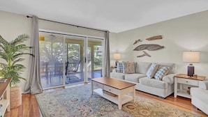 A living room with a sofa, a coffee table, a side table, a lamp, and a large window with curtains. - 3C Mariners Walk by Avantstay Beach & Pool Access, Enclosed Porch (Isle of Palms)