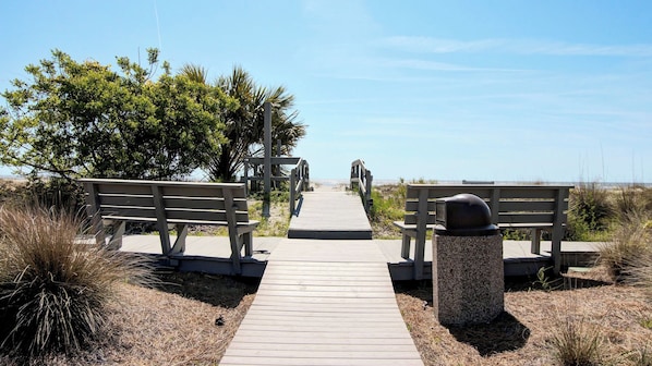 A wooden boardwalk leading to a beach with benches and a trash can.