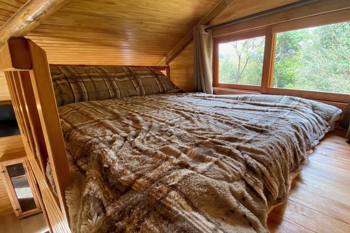 Ein gemütliches Schlafzimmer unter dem Dach, mit holzverkleideter geneigter Decke, einem großen Bett mit flauschiger Decke und einem Fenster mit Blick auf Grünflächen.