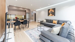 A modern living room with a grey sofa, a dining table with chairs, and a kitchen in the background.
