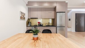A modern kitchen with light-colored cabinets, a stainless steel refrigerator, and a wooden dining table with a potted plant.