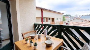 Une table en bois avec des chaises sur un balcon, des plantes en pot et, en arrière-plan, une vue sur des bâtiments et un tuyau rouge.