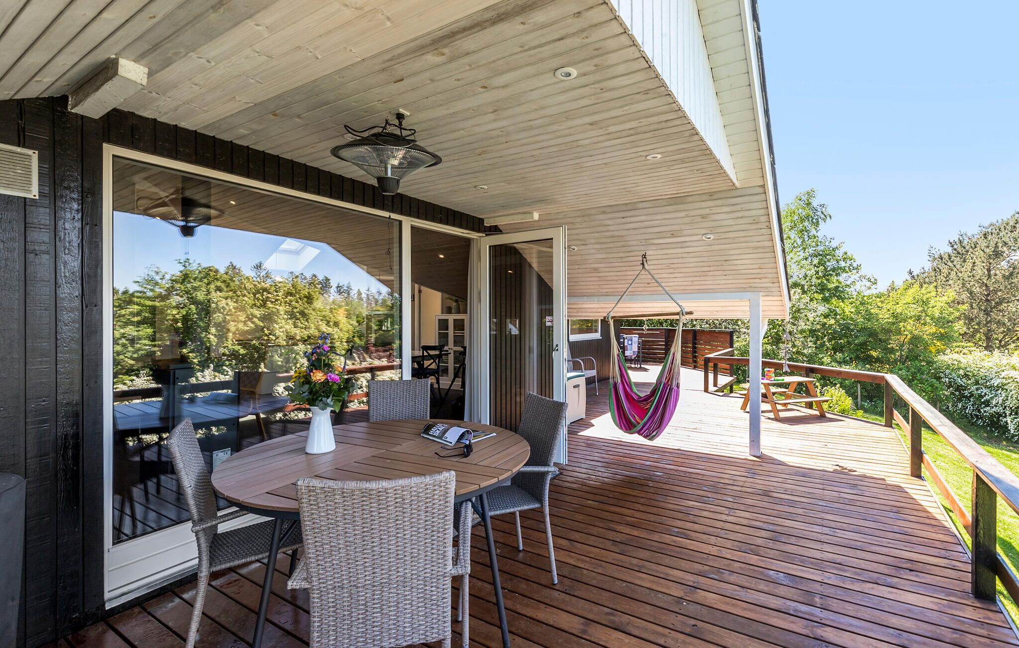 Une terrasse en bois avec une table ronde et des chaises, un hamac et une porte vitrée donnant sur un espace extérieur.