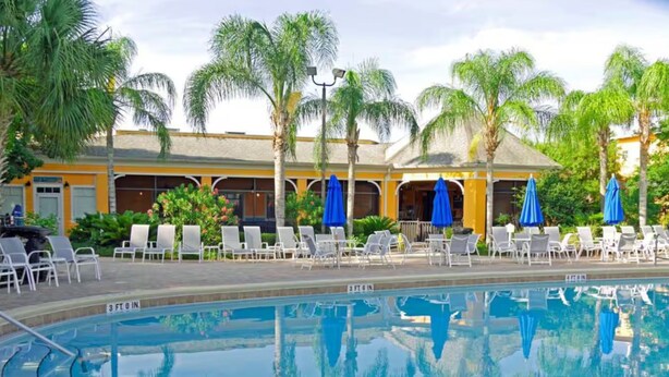 A pool area with lounge chairs and umbrellas, surrounded by palm trees and a building with yellow walls. - Family Friendly Condo near Disney (Davenport)