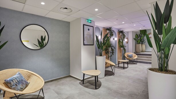 A modern lobby with a grey wall, a round mirror, and several seating areas with plants.