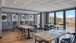 A modern dining area with wooden tables and chairs, framed artwork on the walls, and large windows offering a view of the city.