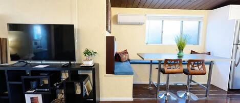 A compact living space with a flat-screen TV, a dining table with chairs, a small kitchen area, and a potted plant.