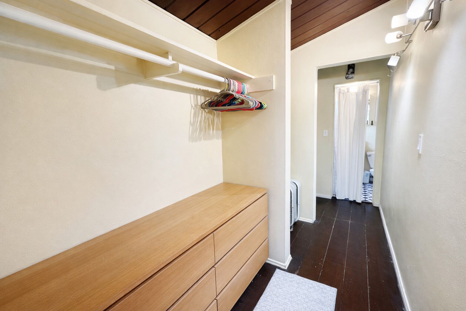 A hallway with a wooden bench, a white wall, a hanging rack with clothes, and a door leading to a bathroom.
