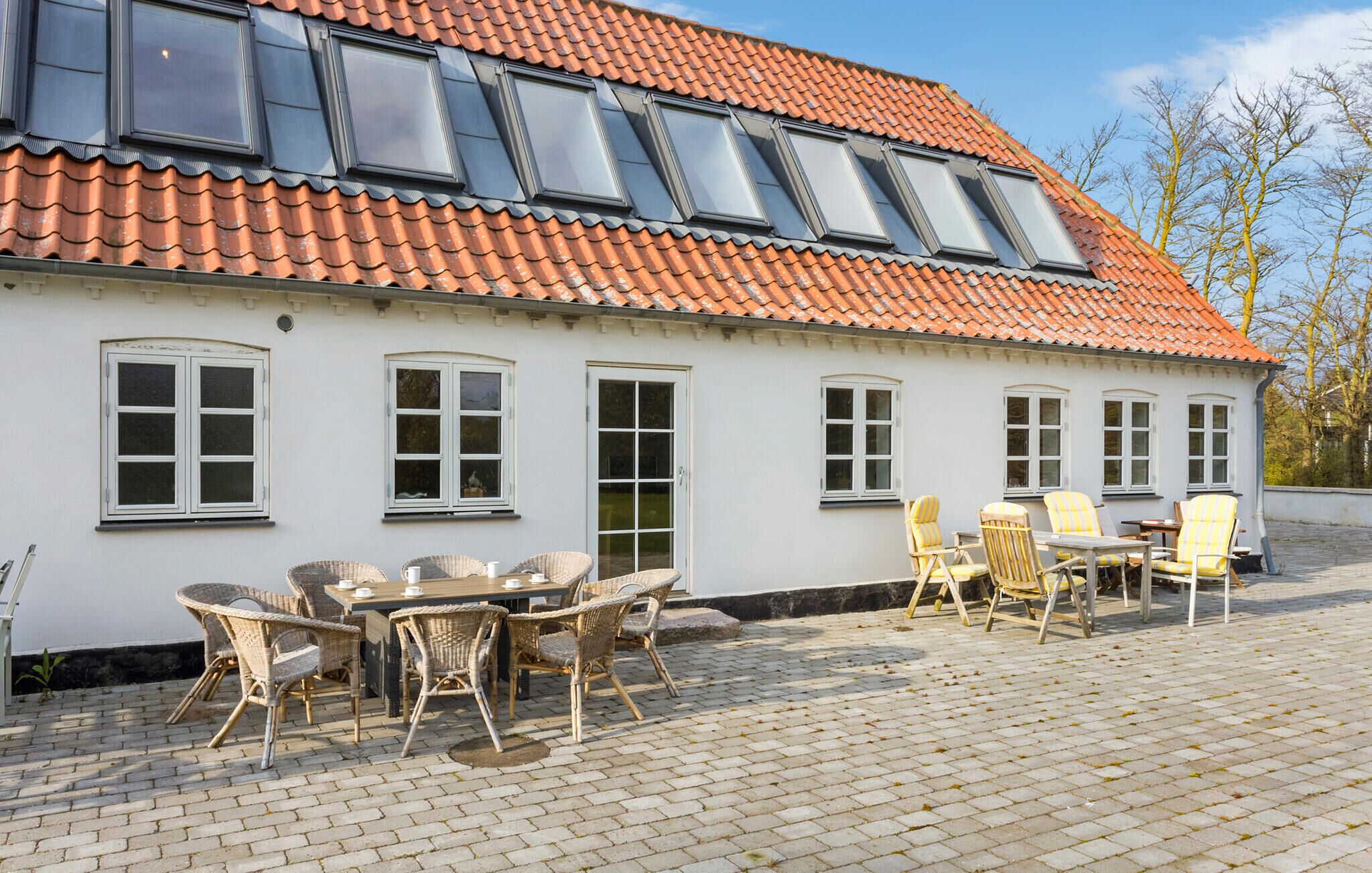 Een wit gebouw met een rode pannendak, meerdere ramen en een geplaveid terras met rieten meubilair.
