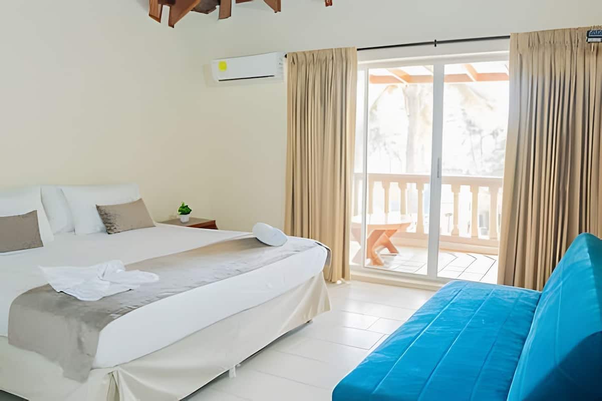 Ein Hotelzimmer mit Bett, blauem Sofa, kleinem Tisch mit Pflanze und einer Schiebetür zum Balkon.