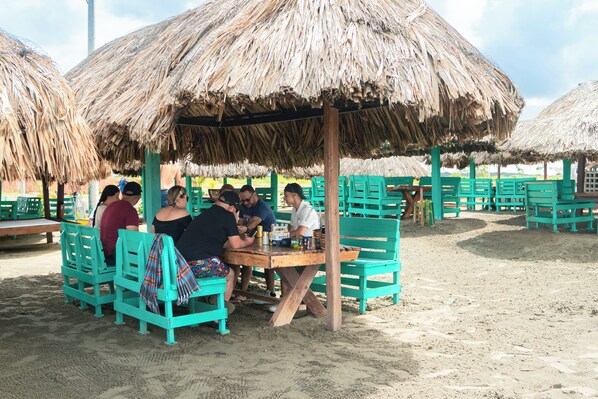 Een zitgedeelte aan het strand met rieten parasols en houten tafels.