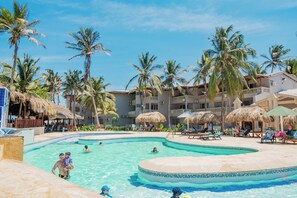 Een zwembadgedeelte van een resort met palmbomen, ligbedden en een gebouw met balkons.