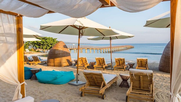 A beachside lounge with wooden lounge chairs, a thatched structure, and umbrellas providing shade.