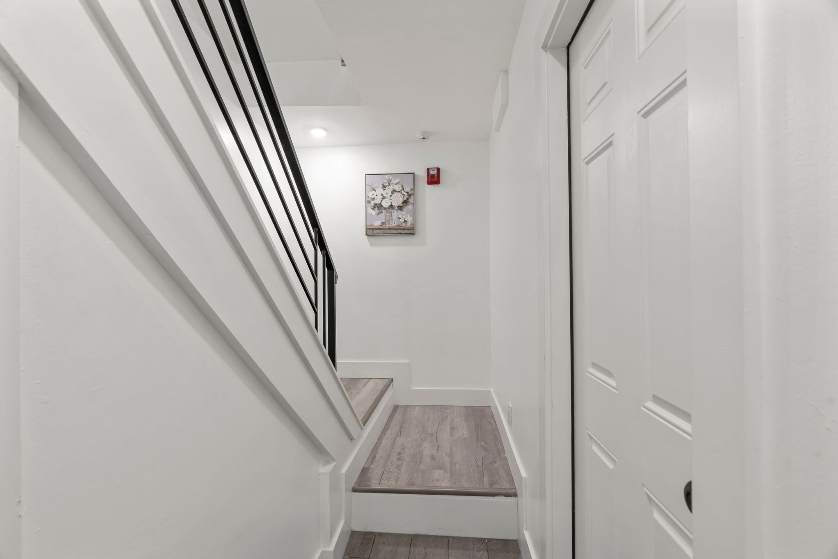 A narrow hallway with white walls, a wooden floor, and a staircase leading to a door.