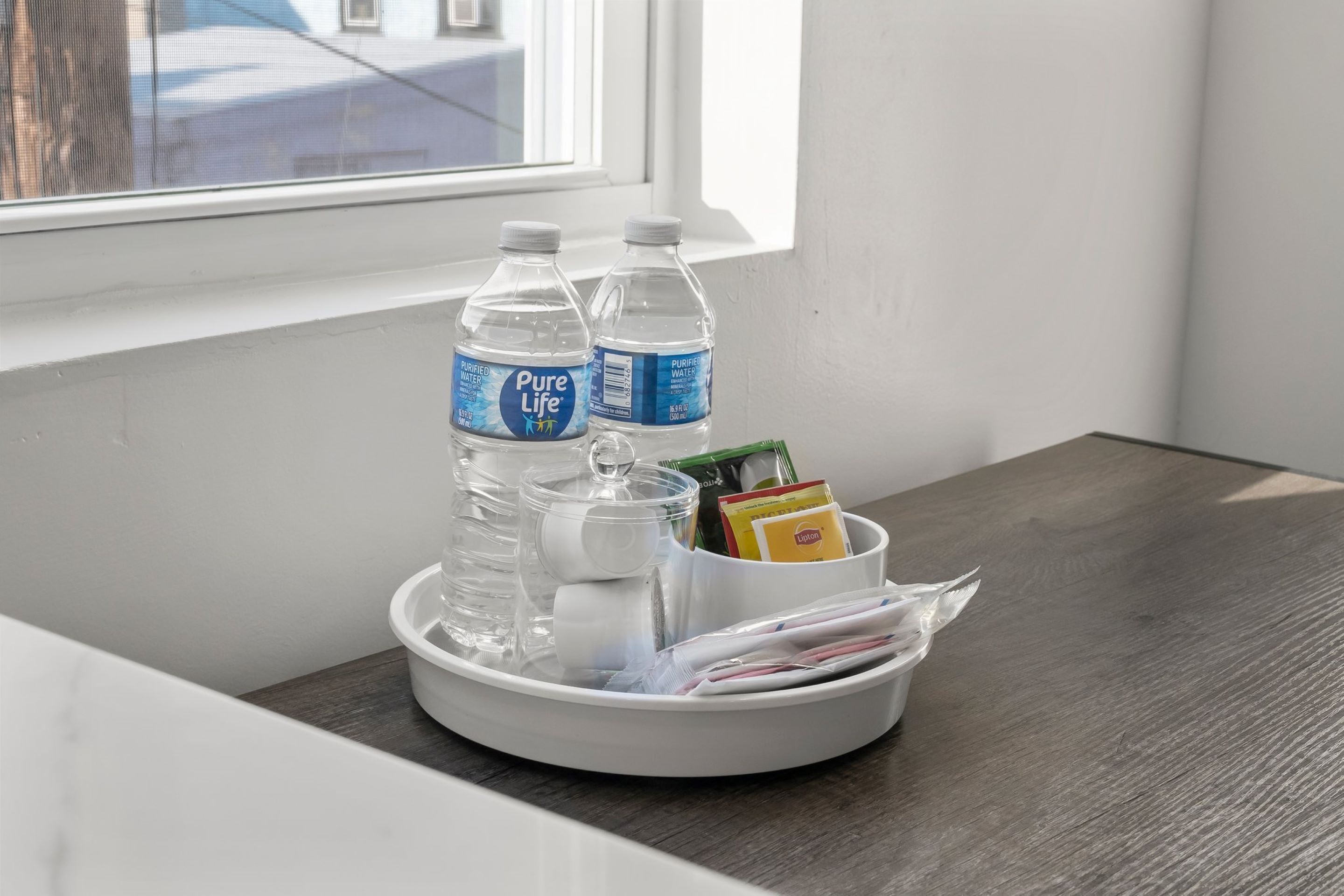 En bakke med to flasker Pure Life vand, et glas, en æske med servietter og nogle magasiner på et træbord nær et vindue.