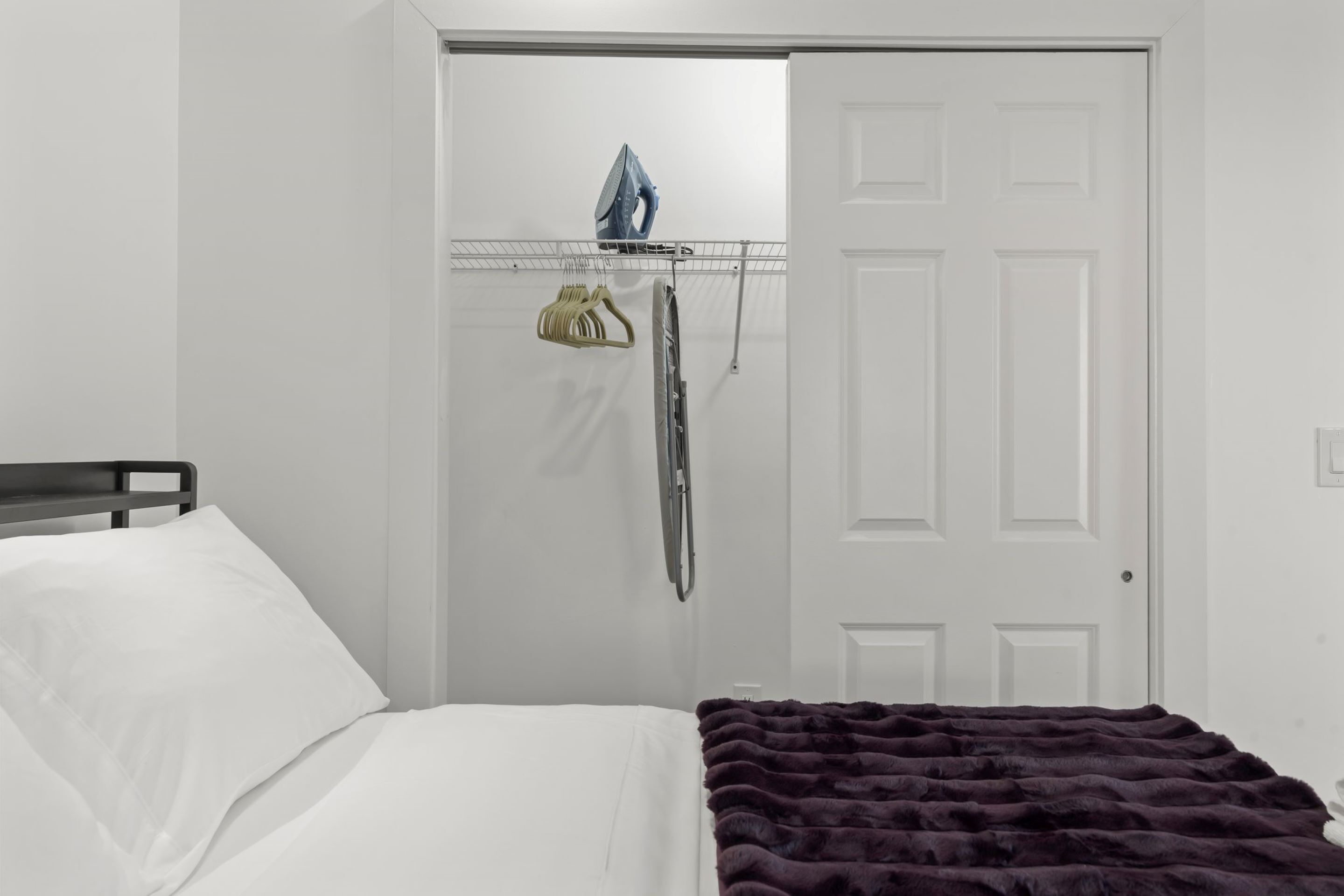 Un dormitorio con cama, una puerta y un tablero de planchar montado en la pared con plancha y perchero.