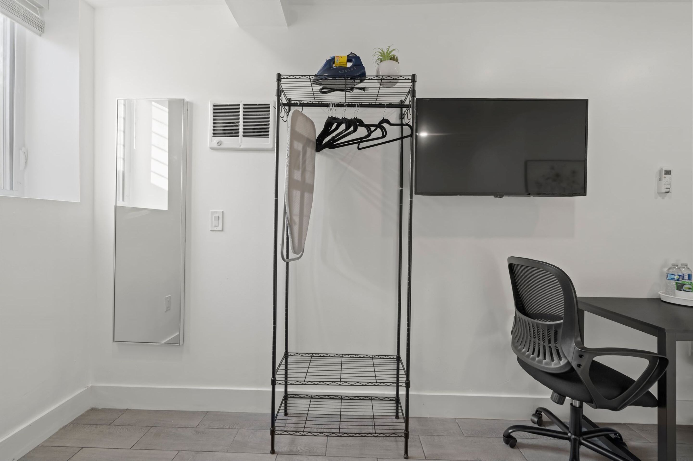 A room with a metal rack holding clothes, a desk with a chair, a TV, and a potted plant.