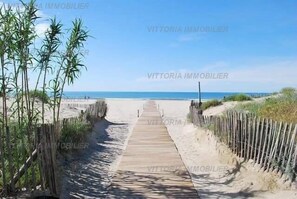 Une promenade en bois mène à une plage bordée de dunes de sable et, de part et d’autre, de plantes de bambou.