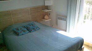 A bedroom with a bed, two patterned pillows, a bedside table, a nightstand, a window with curtains, and a radiator.