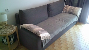 A dark-colored sofa with a patterned throw blanket and a cushion, a small wooden side table with a lamp, and a tiled floor.