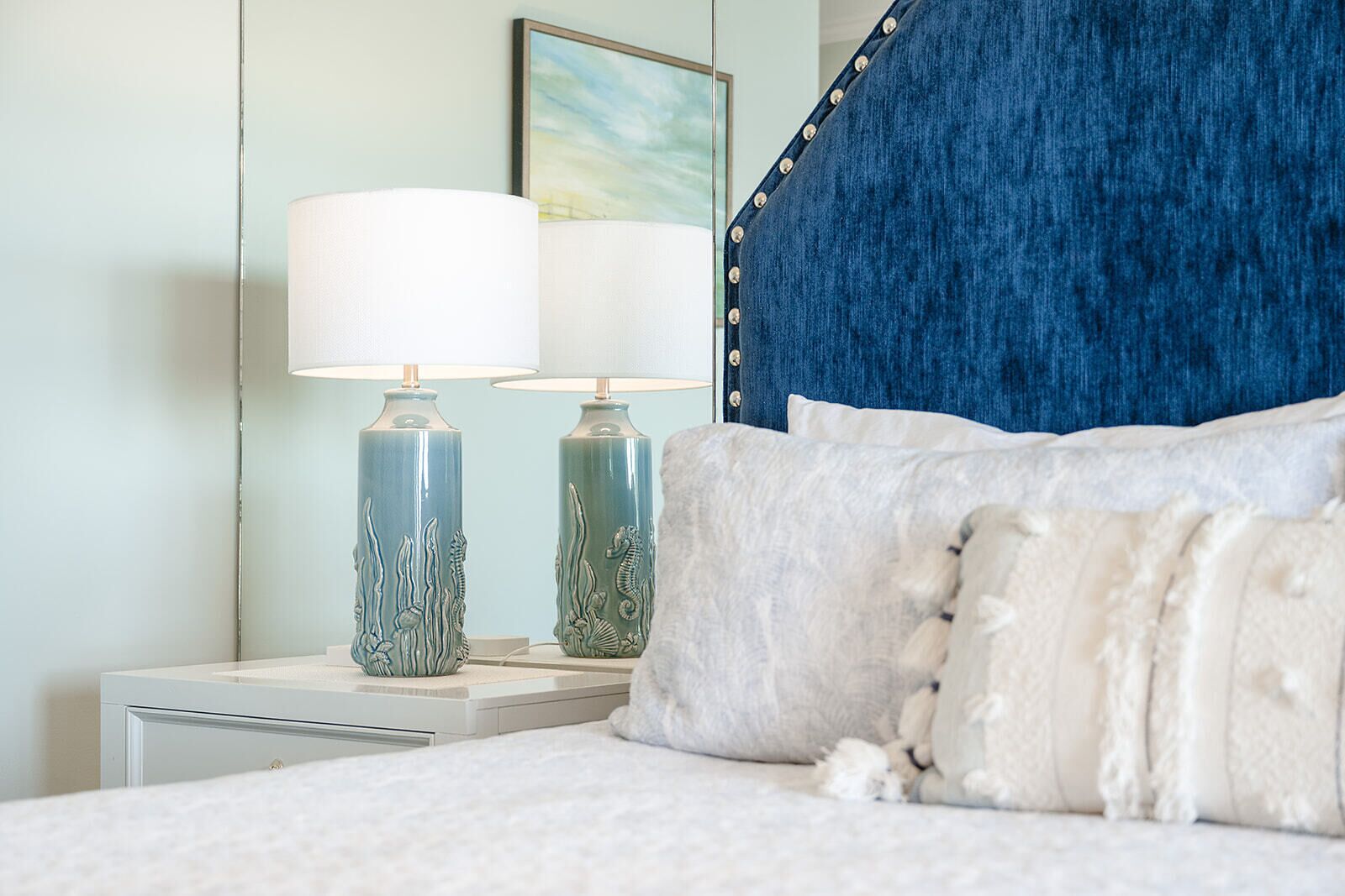 A bed with white bedding, two decorative lamps with sea-themed designs, and a blue upholstered headboard.