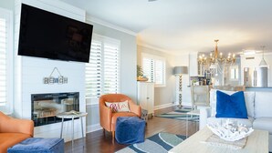 A living room with a fireplace, television, orange armchair, blue ottoman, and a dining area with a chandelier. - Ocean Pearl! Amazing Ocean & Pier Views, Oceanfront Pool, In the heart of the Pier Village! (Saint Simons Island)