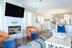 A living room with a fireplace, television, orange armchair, blue ottoman, and a dining area with a chandelier. - Ocean Pearl! Amazing Ocean & Pier Views, Oceanfront Pool, In the heart of the Pier Village! (Saint Simons Island)