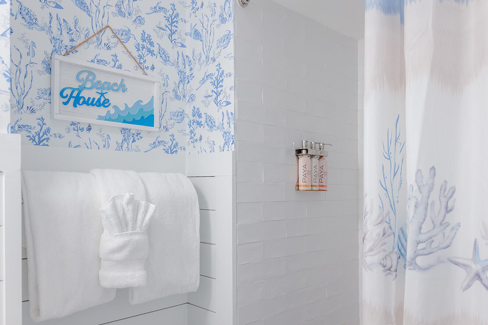 A bathroom with a white tiled wall, a shower curtain with a blue ocean design, a towel rack with a white towel, and a sign that reads 'Beach House'.