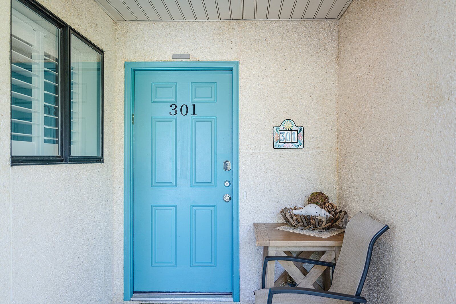 Une porte turquoise portant le numéro 301, une petite table avec un panier, et une plaque fixée au mur, également portant le numéro 301.