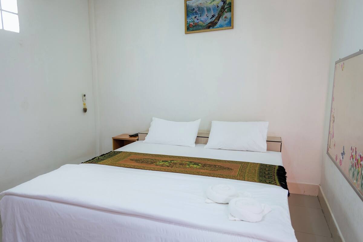 Quarto simples e arejado, com uma cama, mesa de cabeceira, um quadro na parede e um tapete estampado.