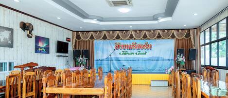 A conference room with wooden tables and chairs, a stage with a banner, and a large window.