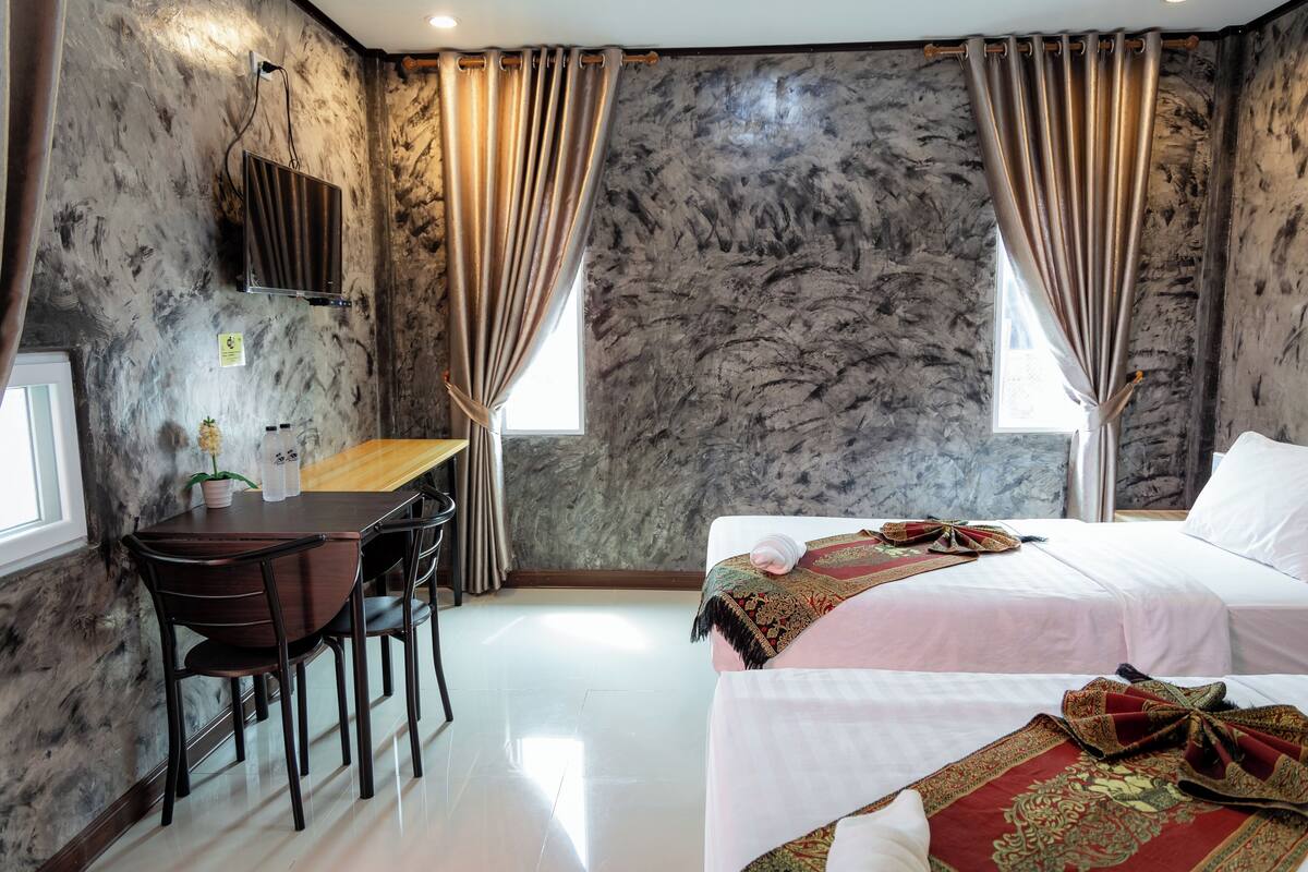 Quarto de hotel com parede de mármore, uma escrivaninha com cadeiras, duas camas com roupa de cama vermelha e branca e uma janela com cortinas.