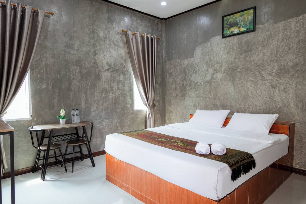Quarto de hotel com uma cama grande, uma mesa pequena com cadeiras, uma janela com cortinas e um quadro na parede.