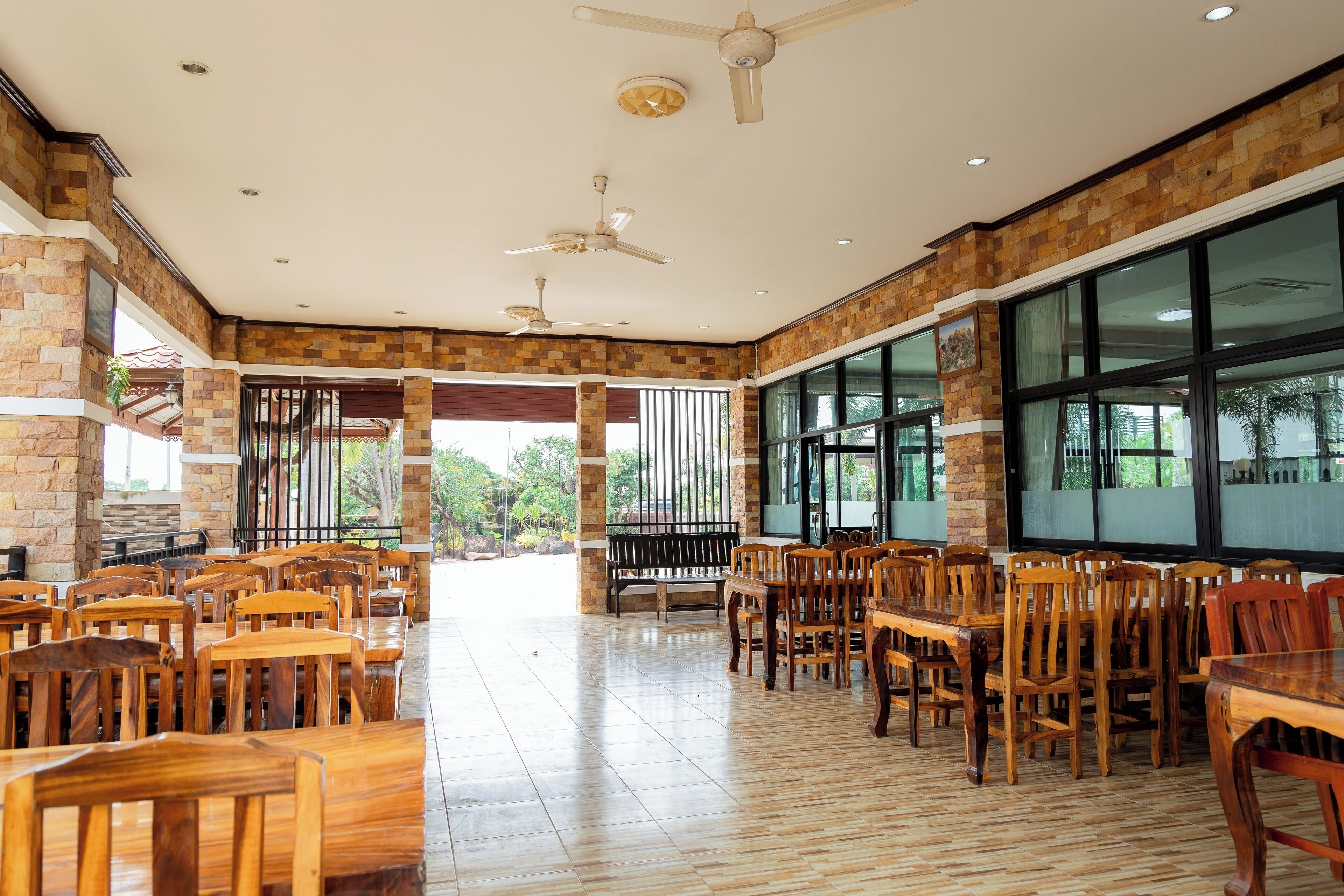 A spacious indoor area with multiple wooden tables and chairs, ceiling fans, and large windows allowing natural light.
