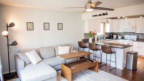 A modern living room with a sofa, coffee table, and a dining area with bar stools. A kitchen with a microwave and island is visible in the background.