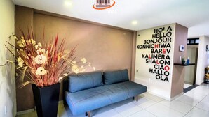 Un moderno vestíbulo de hotel con un sofá gris, una planta decorativa y una pared con saludos en varios idiomas.