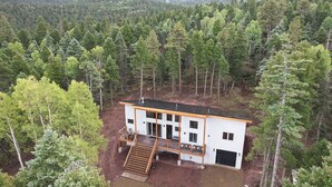 A modern two-story cabin surrounded by dense forest, with a wooden deck and a large sliding glass door entrance. - Alpine View: Your Getaway to Mountain Adventures (Angel Fire)