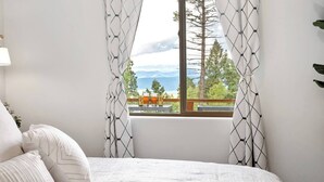 A bedroom with a bed, white curtains, and a view of trees and mountains through a window. - Alpine View: Your Getaway to Mountain Adventures (Angel Fire)
