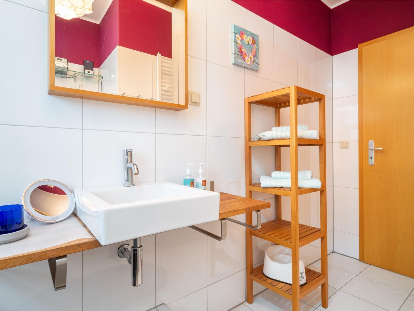 Een badkamer met een witte wastafel, een houten plank met handdoeken, een spiegel en een deur.